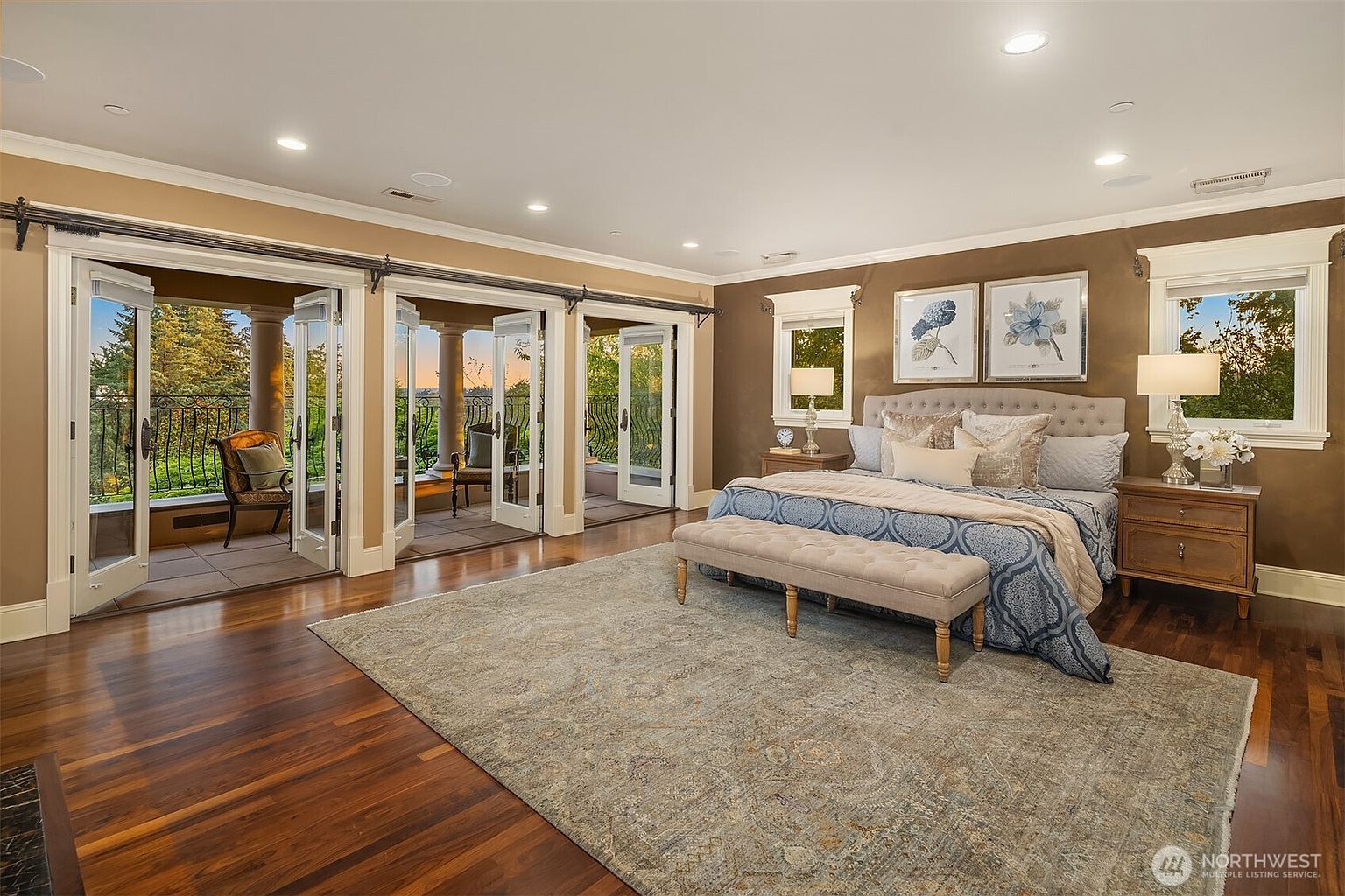 This spacious primary bedroom features rich hardwood flooring, a large area rug, and a king-sized bed with elegant blue patterned bedding. The room is highlighted by a wall of French doors that open onto a private balcony, allowing for abundant natural light and scenic views. The sophisticated decor, including framed artwork and matching bedside lamps, creates a luxurious and tranquil atmosphere.