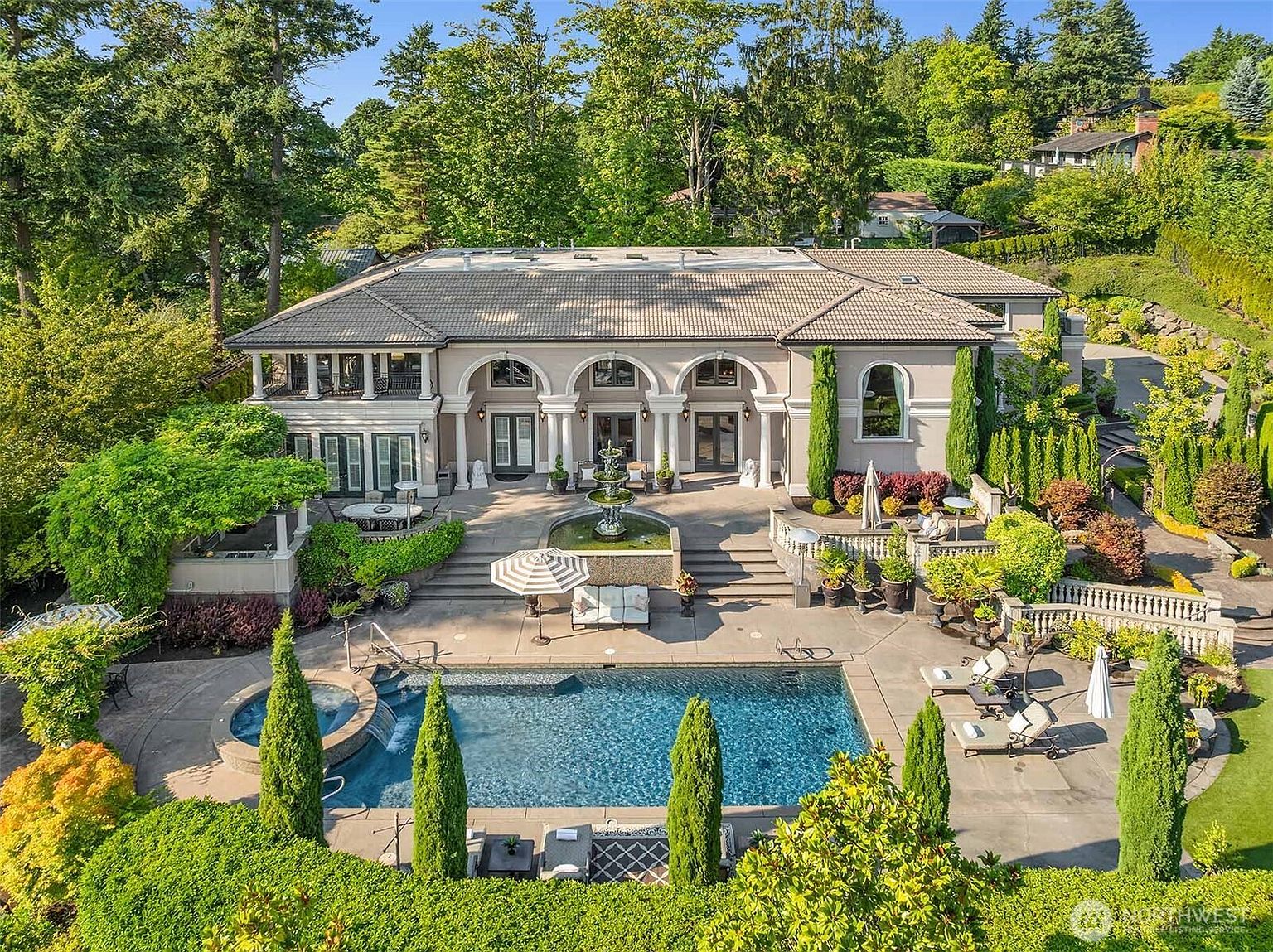 This stunning Mediterranean-style estate features a grand rear facade with arched doorways, stone columns, and a symmetrical architectural design. The expansive outdoor living space includes a tiered patio, a central tiered fountain, and a luxurious swimming pool with an integrated spa, all surrounded by lush, manicured landscaping. The elevated perspective captures the seamless integration of high-end residential architecture with a resort-like backyard oasis.