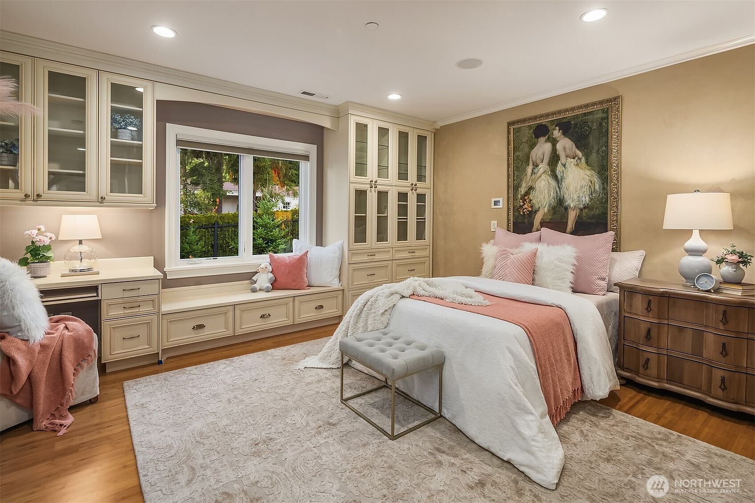 This charming, well-lit bedroom features a soft, feminine color palette with blush pink accents and cream-colored built-in cabinetry. The room includes a cozy window seat, a desk area, and a bed adorned with plush pillows, all set against a warm, neutral wall color. The perspective is a wide-angle shot that captures the functional layout and elegant, inviting atmosphere of the space.