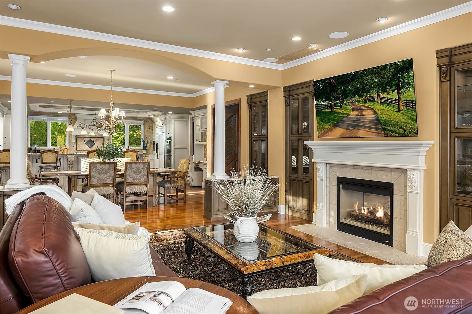 This elegant living room features a warm, inviting atmosphere with a large leather sofa, a glass-topped coffee table, and a prominent fireplace with a mounted television above it. The open-concept layout seamlessly transitions into a formal dining area, highlighted by classic columns and rich hardwood flooring. The space is bathed in soft, recessed lighting, creating a sophisticated and comfortable environment perfect for entertaining.