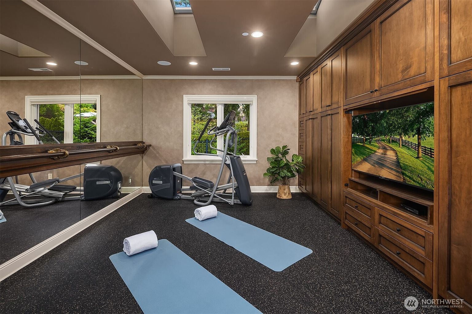 This well-appointed home gym features a sleek, dark rubberized floor, a large wall-to-wall mirror with a wooden ballet barre, and an elliptical machine. The room is accented by custom built-in wooden cabinetry that houses a large television, creating a functional and sophisticated workout space. Natural light streams in through a window and overhead skylights, enhancing the bright and inviting atmosphere.