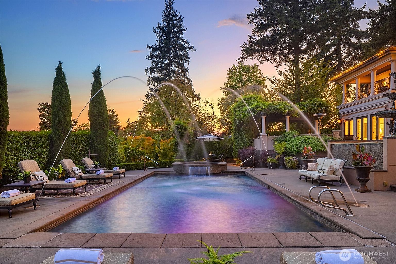 This stunning backyard oasis features a rectangular swimming pool with integrated water fountains and a raised circular spa, creating a luxurious resort-like atmosphere. The surrounding patio is furnished with elegant lounge chairs and outdoor seating, all set against a backdrop of lush greenery and a beautiful sunset sky. The perspective is a low-angle, eye-level shot that emphasizes the grandeur of the pool area and the sophisticated architectural details of the adjacent home.