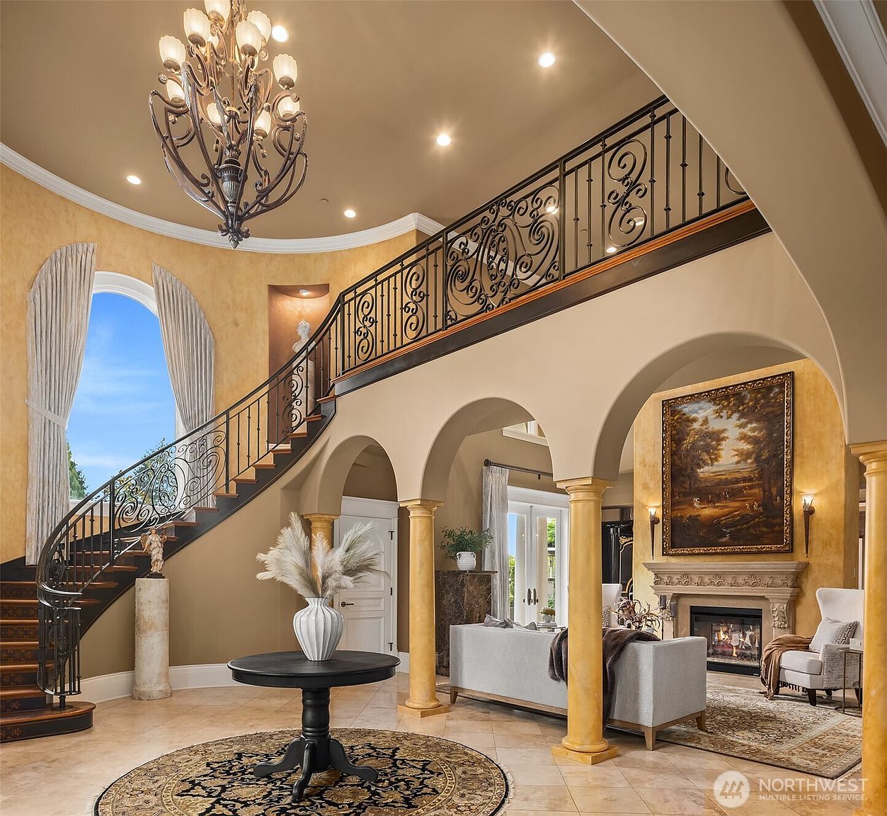 This grand foyer features a dramatic curved staircase with intricate wrought-iron railings, leading to an upper-level landing. The space is defined by soaring ceilings, elegant arched columns, and a sophisticated chandelier, creating a luxurious and welcoming atmosphere. A circular table with a decorative vase sits atop a patterned rug, while a cozy seating area with a fireplace is visible through the arched entryway.