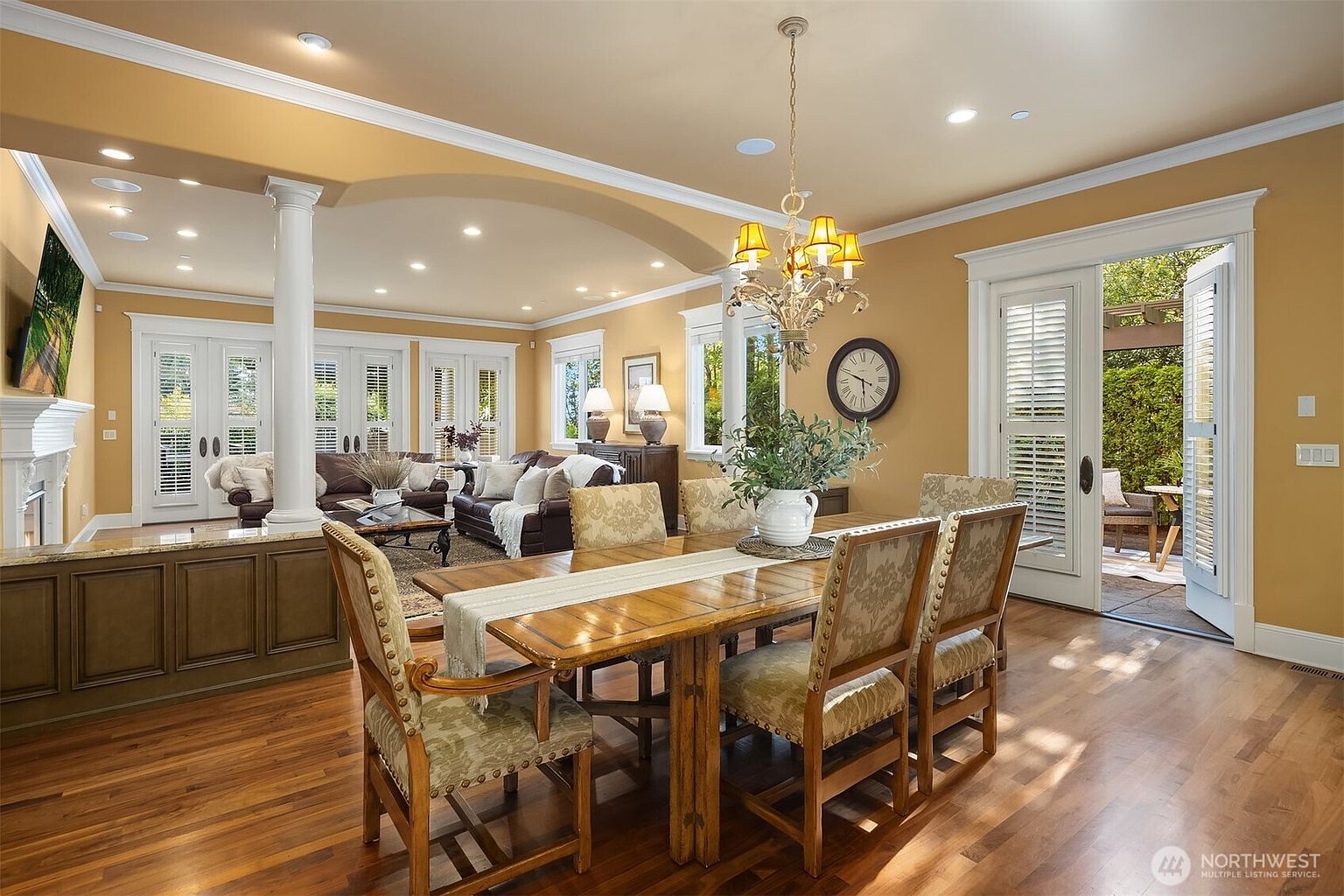 This elegant dining room features a warm, traditional aesthetic with a rich wooden table and upholstered chairs centered under a classic chandelier. The space flows seamlessly into an adjacent living area, separated by architectural columns and a low-profile built-in cabinet. Large glass doors open to a lush outdoor patio, bathing the room in natural light and creating an inviting, open-concept atmosphere.