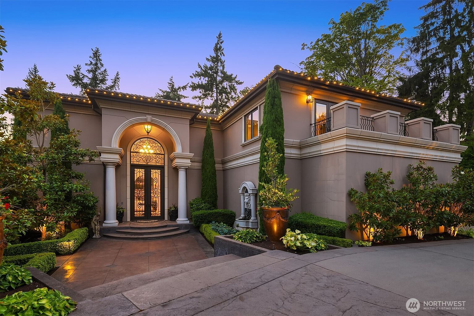 This elegant, Mediterranean-inspired estate features a grand arched entryway with ornate double doors and classic columns, set against a warm, dusk-lit sky. The exterior is characterized by smooth stucco walls, decorative roofline lighting, and a manicured landscape with tall cypress trees and a stone fountain. The perspective is a low-angle, wide shot that emphasizes the home's architectural sophistication and welcoming curb appeal.
