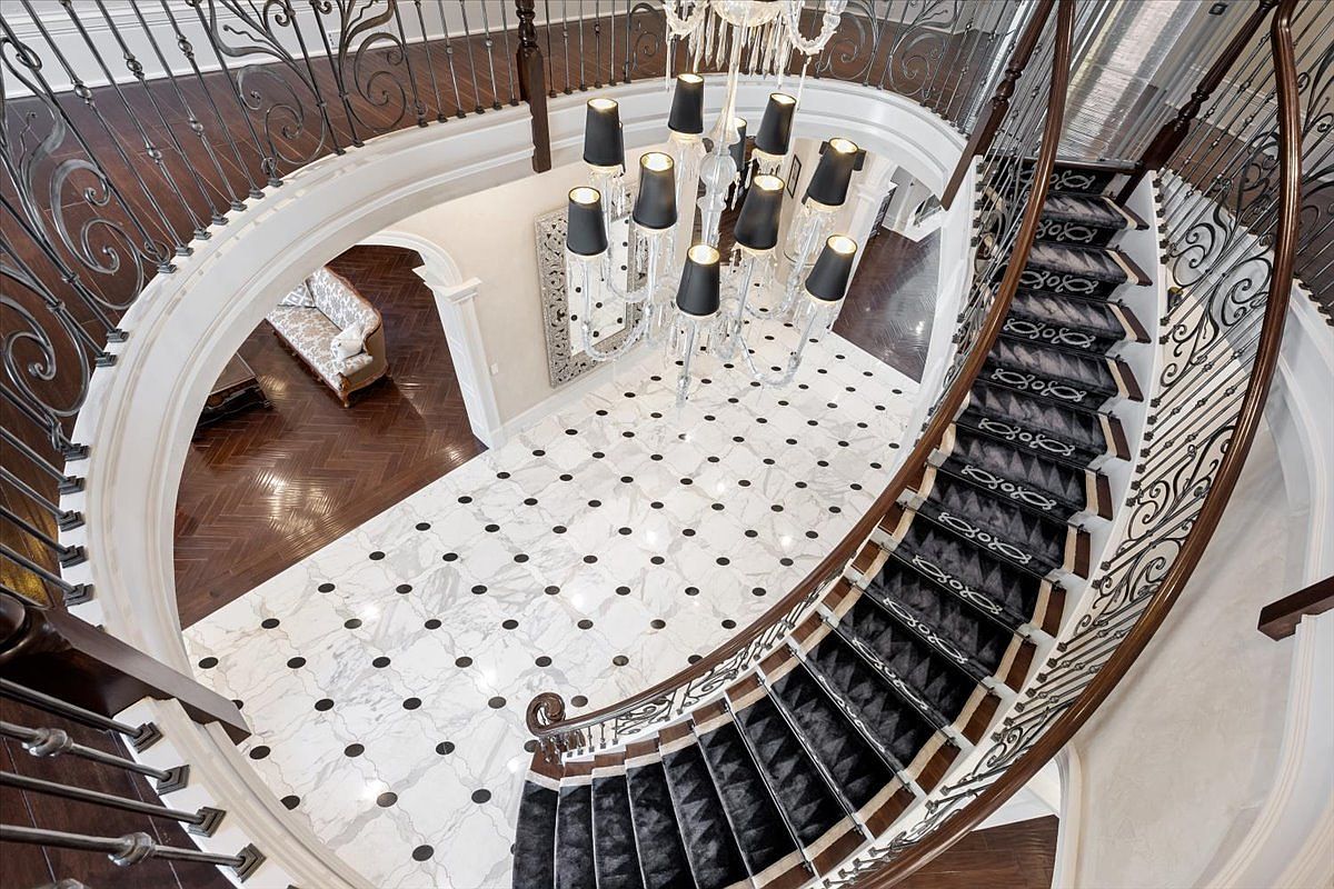 This high-angle, cinematic shot captures a grand foyer featuring a sweeping curved staircase with ornate metal railings and dark-toned treads. The floor is finished in light-colored stone with a repeating dark circular pattern, centered beneath a large, multi-tiered crystal chandelier. To the side, an arched entryway reveals a glimpse of a formal living area with dark wood flooring and a classic sofa, creating an impression of luxury and architectural elegance.