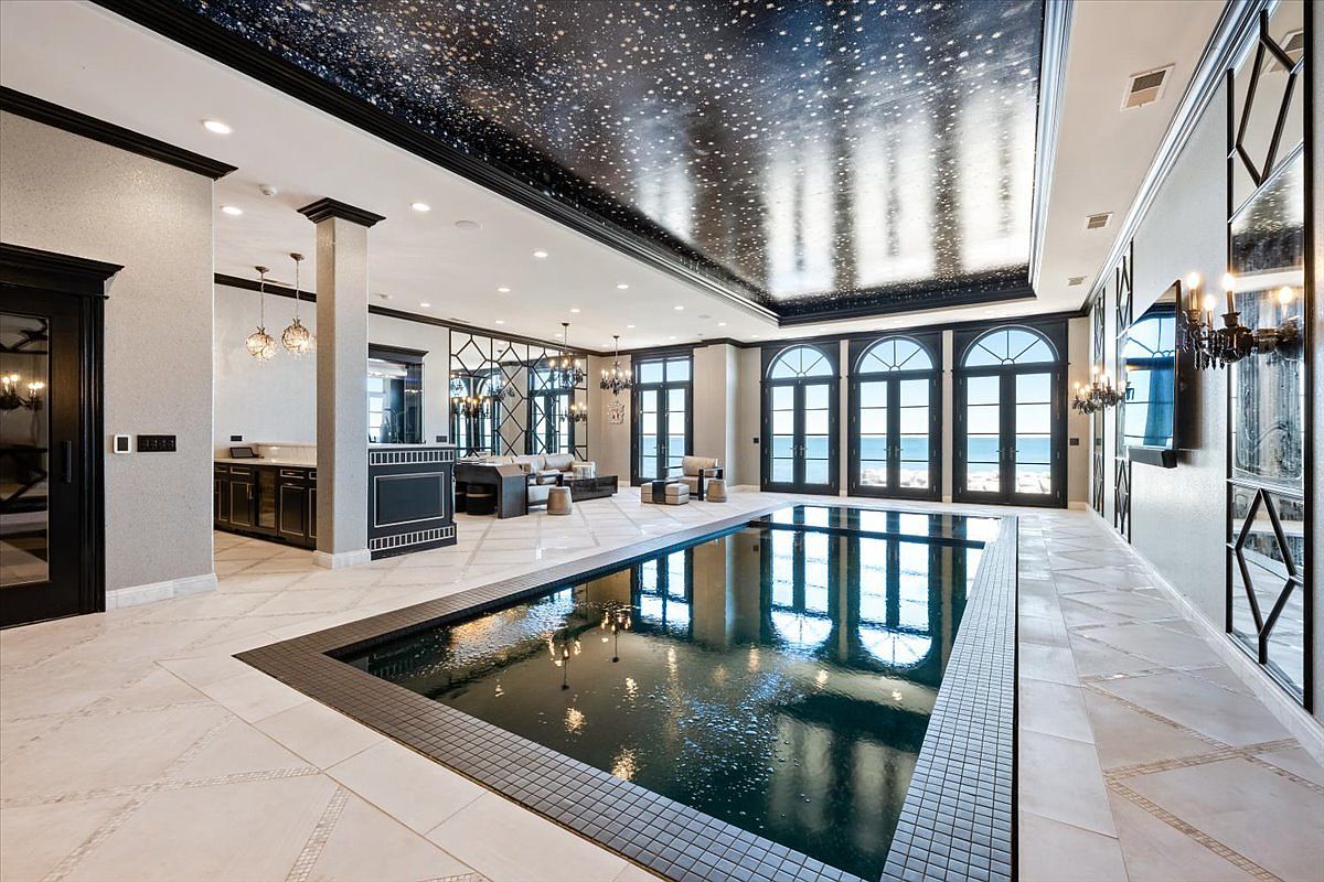 This luxurious indoor pool room features a dramatic ceiling painted to resemble a starry night sky, reflecting beautifully in the dark-tiled pool below. The space is framed by large, arched windows that provide a view of the outdoors, while mirrored wall panels and elegant light fixtures enhance the sophisticated, high-end atmosphere. A seating area is visible in the background, creating a seamless transition between the aquatic space and a lounge area.