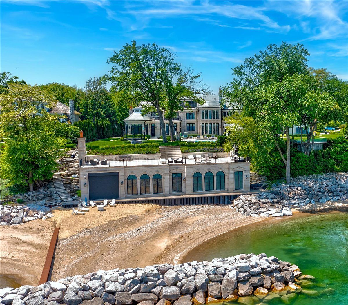 This elevated perspective captures a luxurious waterfront estate featuring a grand multi-story residence set back from the shore, with a sophisticated stone-clad boathouse structure positioned directly on the beach. The property boasts extensive tiered landscaping, a spacious stone terrace atop the boathouse, and a private sandy beach protected by a rock breakwater. The scene conveys an atmosphere of exclusive coastal living, highlighting the seamless integration between the refined architecture and the natural lakeside environment.