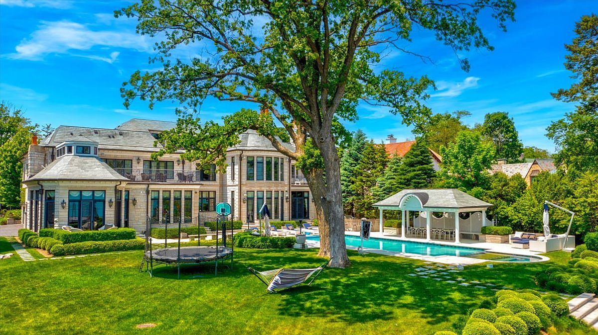 This expansive rear view of a luxury estate showcases a multi-story stone residence with a classic architectural style, featuring numerous large windows and a prominent turret-like structure. The meticulously landscaped backyard includes a sprawling green lawn, a large swimming pool with an adjacent covered pavilion, and a mature tree providing shade. A trampoline and a hammock are positioned on the grass, emphasizing the property's suitability for outdoor recreation and leisure.