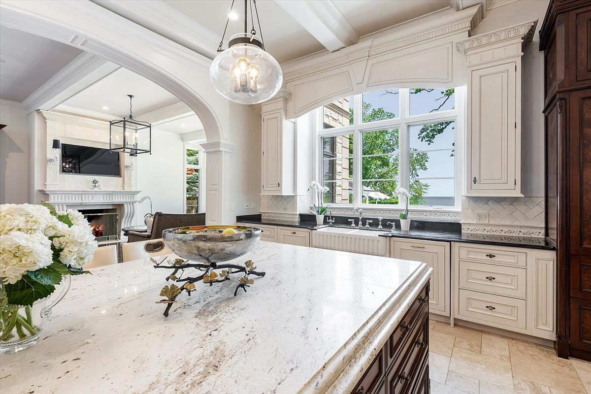 This elegant kitchen features a large central island with a light-colored stone countertop and a decorative metal bowl centerpiece. The space is anchored by white cabinetry, a farmhouse-style sink positioned beneath a large window, and dark wood accents that provide a sophisticated contrast. An arched opening leads into an adjacent living area, creating an open and airy flow that highlights the home's refined architectural details.