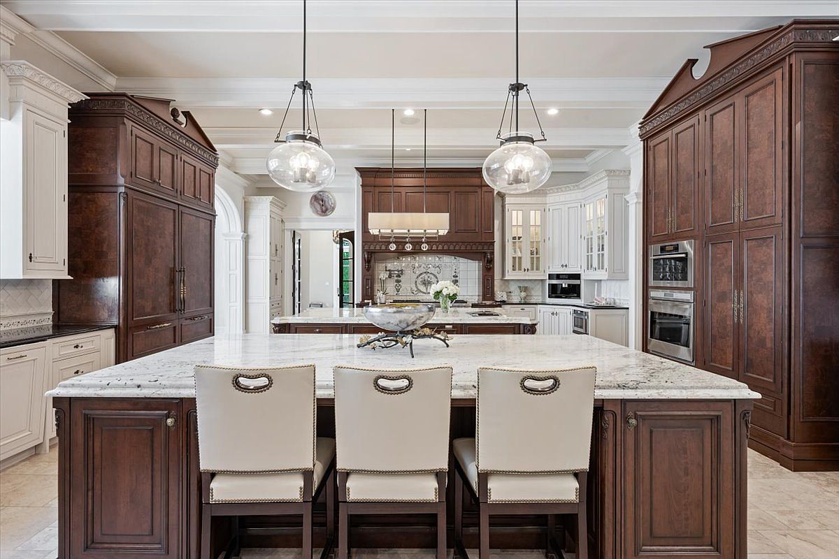 This expansive, high-end kitchen features a large central island with seating for three, topped with a light-colored stone surface. The space is defined by rich, dark wood cabinetry, integrated appliances, and elegant pendant lighting that hangs over the island. The perspective is centered and symmetrical, showcasing a sophisticated design with a secondary island in the background and a neutral tile floor throughout.