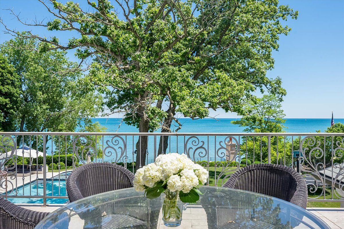 This elevated balcony offers a serene, panoramic view of a large body of water framed by a mature, leafy tree. In the foreground, a glass-topped table holds a vibrant bouquet of white hydrangeas, flanked by two dark wicker chairs. Below the decorative metal railing, a glimpse of a swimming pool and lush landscaping suggests a luxurious, private outdoor retreat.