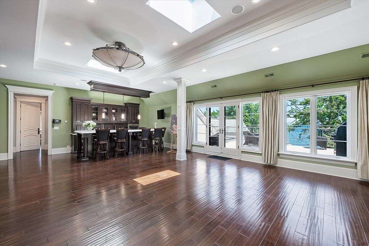 This spacious, open-concept kitchen and living area features dark wood flooring and sage green walls, creating a warm and inviting atmosphere. A large central island with bar seating serves as a focal point, complemented by dark cabinetry and a decorative light fixture hanging from a tray ceiling with a skylight. Large glass doors and windows provide ample natural light and a view of the outdoor deck, enhancing the airy feel of the room.