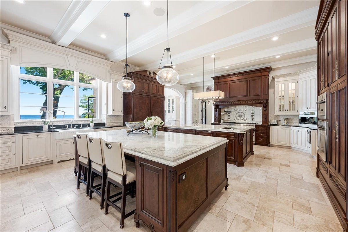 This expansive, high-end kitchen features a dual-island layout with dark wood cabinetry and light-toned stone countertops. Large windows provide a scenic view of the water, while elegant pendant lights hang above the primary island, which includes seating for three. The space is finished with light-colored tile flooring and a decorative tiled backsplash behind the range, creating a sophisticated and airy atmosphere.