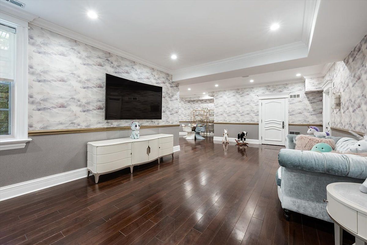 This spacious basement living area features dark hardwood flooring and walls adorned with a soft, cloud-patterned wallpaper above a neutral wainscoting. A large flat-screen television is mounted above a long, white media console, while a plush, light-colored sofa provides comfortable seating in the foreground. The room is illuminated by recessed lighting and includes a glimpse of a dining area in the background, creating a versatile and inviting space.