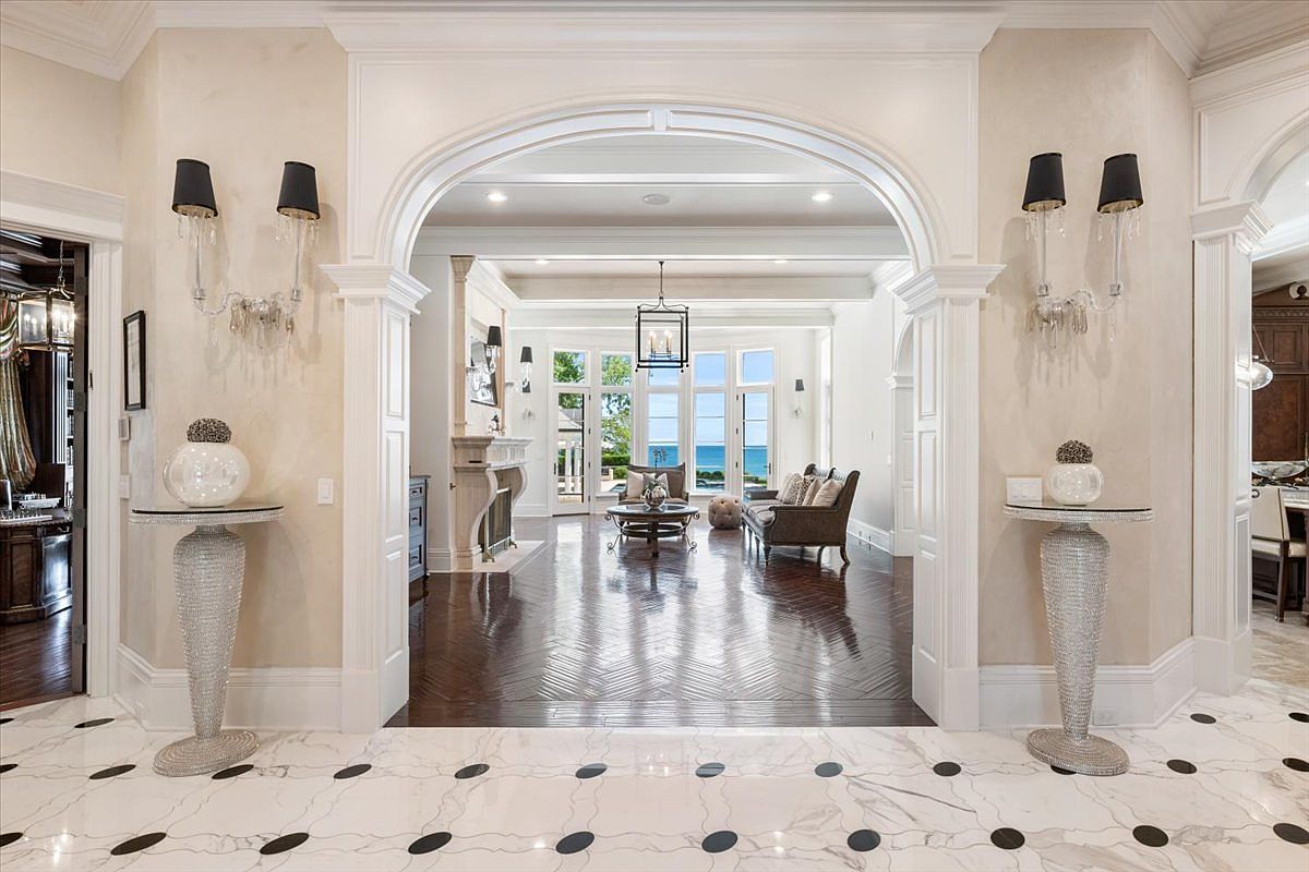 This elegant entryway features a grand arched opening that leads into a bright living area with views of the water. The foreground showcases a white stone floor with dark circular inlays, flanked by two ornate, textured pedestals topped with decorative spheres and wall-mounted sconces. The transition into the living space reveals dark wood flooring arranged in a herringbone pattern, creating a sophisticated contrast between the foyer and the main living area.