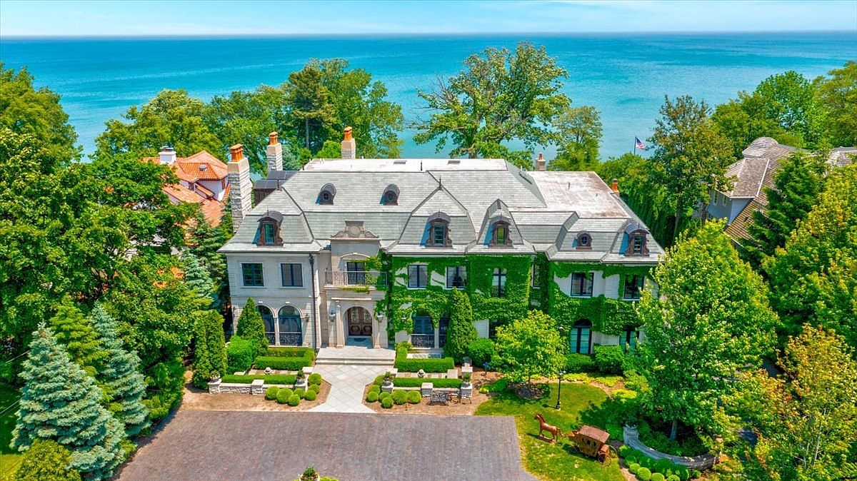 This aerial perspective captures a grand, multi-story estate featuring classic French-inspired architecture with a steep, slate-tiled roof and dormer windows. The exterior is partially adorned with lush climbing ivy, set against a backdrop of mature trees and a vast, serene body of water. A paved driveway leads to the main entrance, complemented by manicured landscaping and stone garden walls.