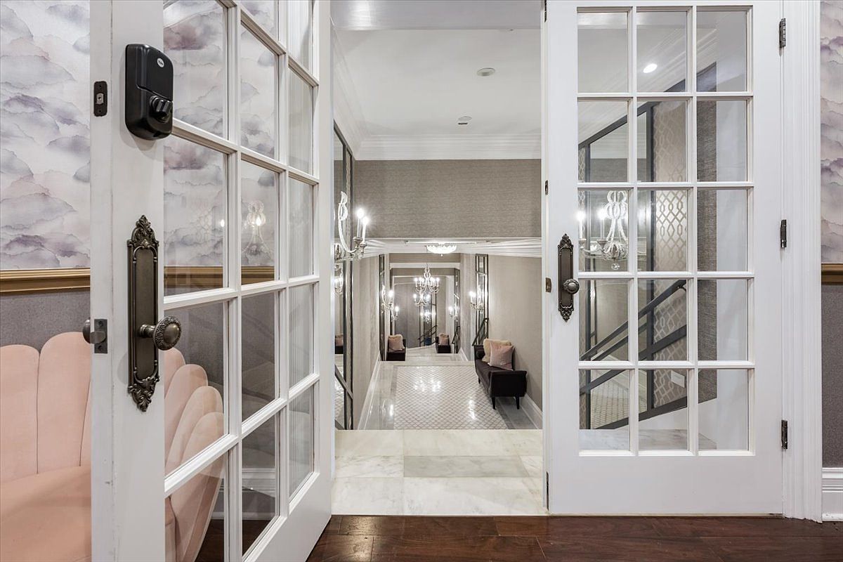 This perspective looks through a set of white French doors with glass panes into a grand, symmetrical hallway. The space features a polished stone floor, mirrored wall panels, and elegant chandeliers that create a sense of depth and luxury. A dark bench and soft lighting accents contribute to the sophisticated, formal atmosphere of the entryway.