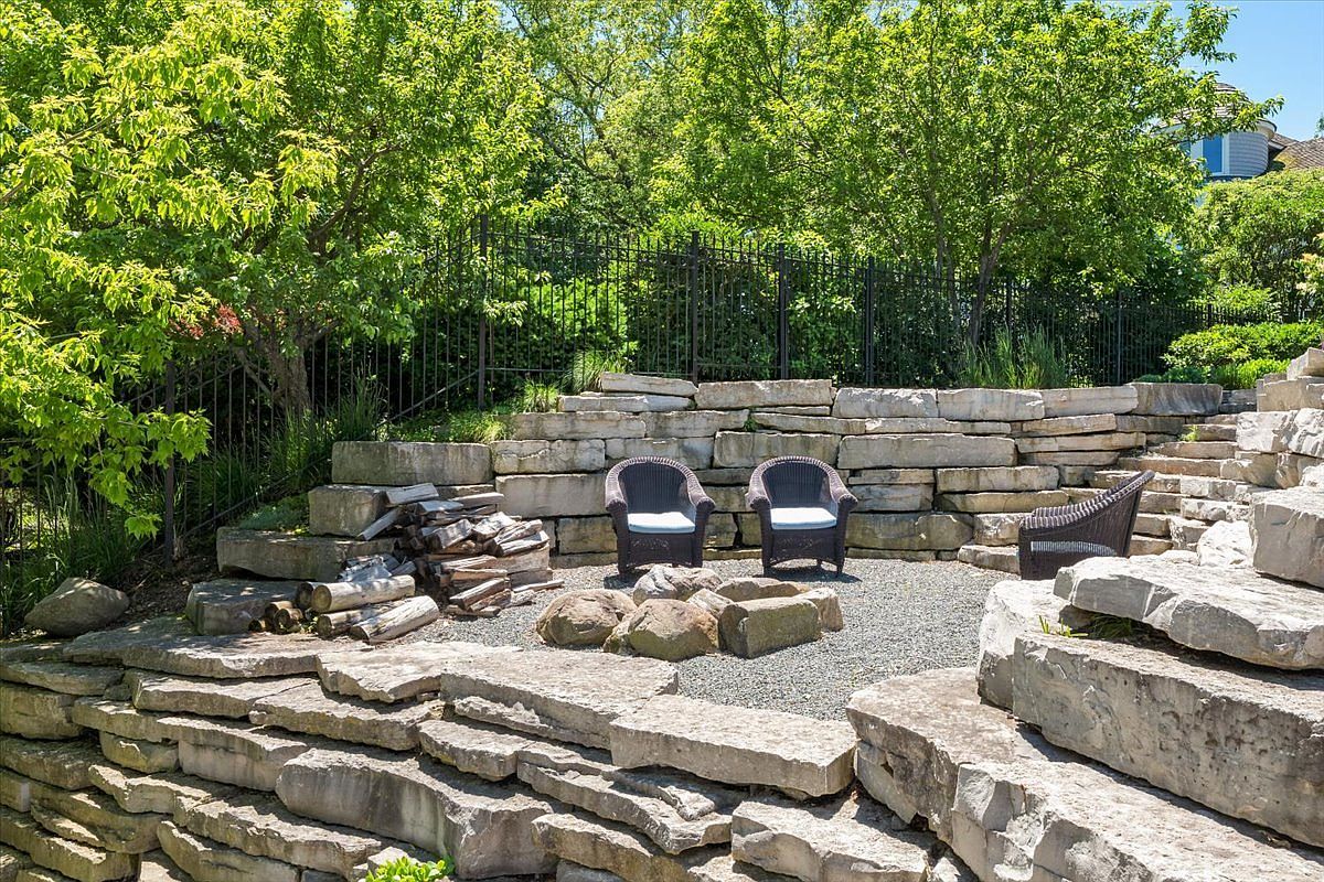 This outdoor space features a tiered landscape design constructed with large, natural stone retaining walls that create a rustic, terraced effect. A gravel-covered seating area is centered in the lower tier, furnished with dark wicker chairs and a small fire pit arrangement. Lush green trees and a black metal fence provide a private, serene backdrop for this well-maintained garden retreat.
