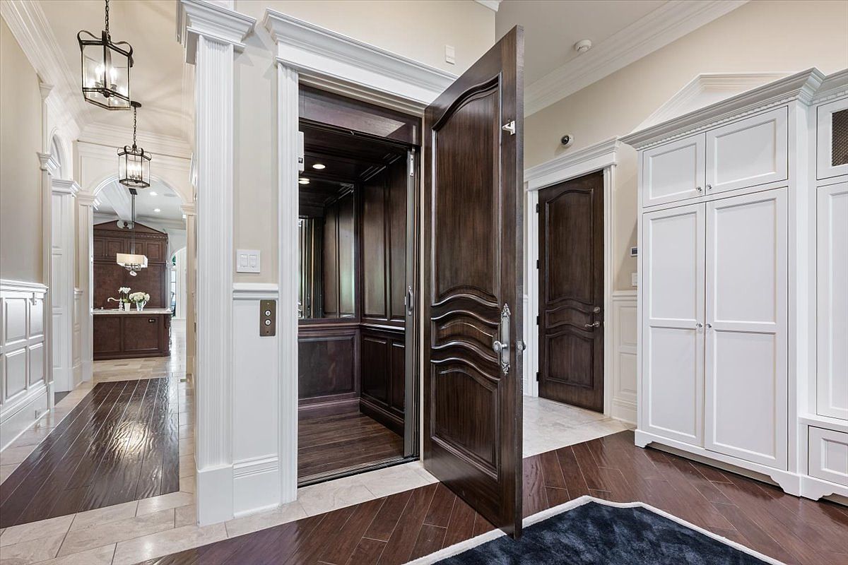 This interior view captures a sophisticated hallway featuring a private elevator with dark wood paneling and a matching ornate door. To the right, a large white built-in storage unit provides ample organization, while a glimpse into an adjacent room reveals a kitchen area with dark cabinetry and pendant lighting. The space is finished with polished dark wood flooring and elegant trim work, creating a luxurious and cohesive aesthetic.