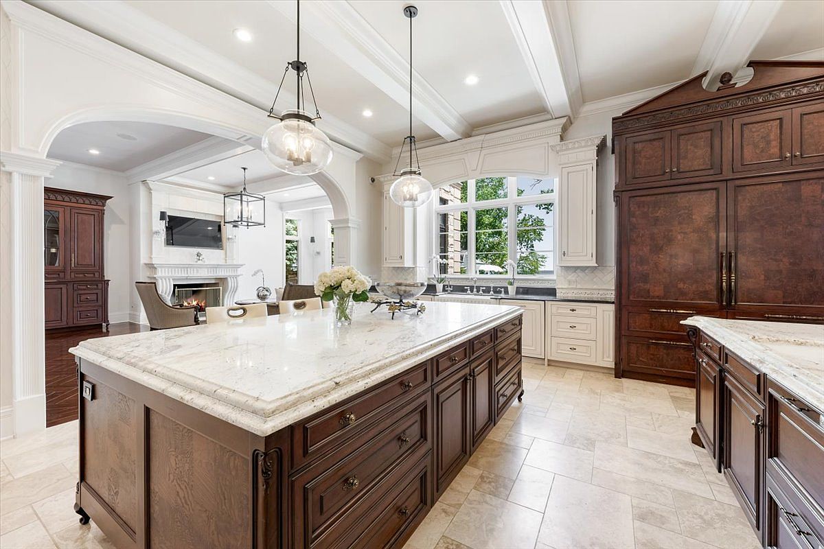 This expansive, high-end kitchen features a large central island with a light-toned stone countertop and dark wood cabinetry. The space is illuminated by two glass pendant lights and recessed lighting, with an open transition leading into a living area that showcases a fireplace and a television. Large windows above the sink area provide ample natural light, while the overall design emphasizes a sophisticated, traditional aesthetic with high-quality finishes.