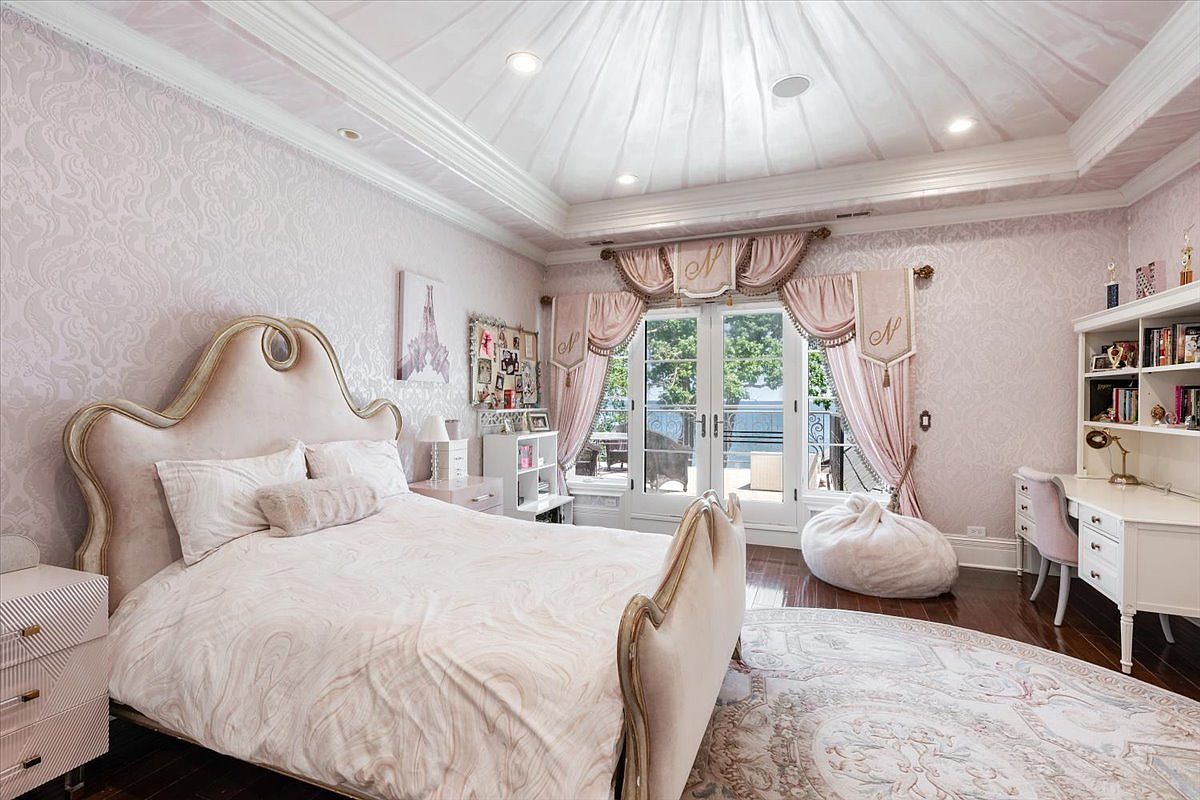 This elegant bedroom features a soft, monochromatic pink color palette with intricate damask wallpaper and a matching upholstered bed frame. The room is anchored by a large, ornate area rug on dark wood flooring and includes a dedicated study area with a white desk and built-in shelving. Large glass doors with custom drapery provide natural light and a view of the outdoor balcony and water beyond.