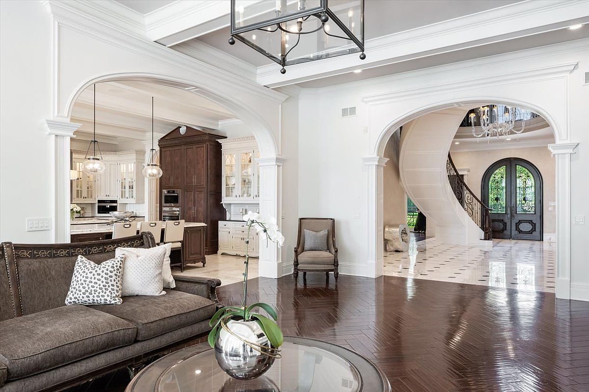 This elegant living area features dark, herringbone-patterned wood flooring and a sophisticated neutral color palette. The space is defined by grand, arched architectural openings that lead into a bright kitchen on the left and a dramatic foyer with a curved staircase on the right. A plush sofa, a glass-topped coffee table, and a single accent chair create a refined seating arrangement under a large, dark-framed lantern chandelier.