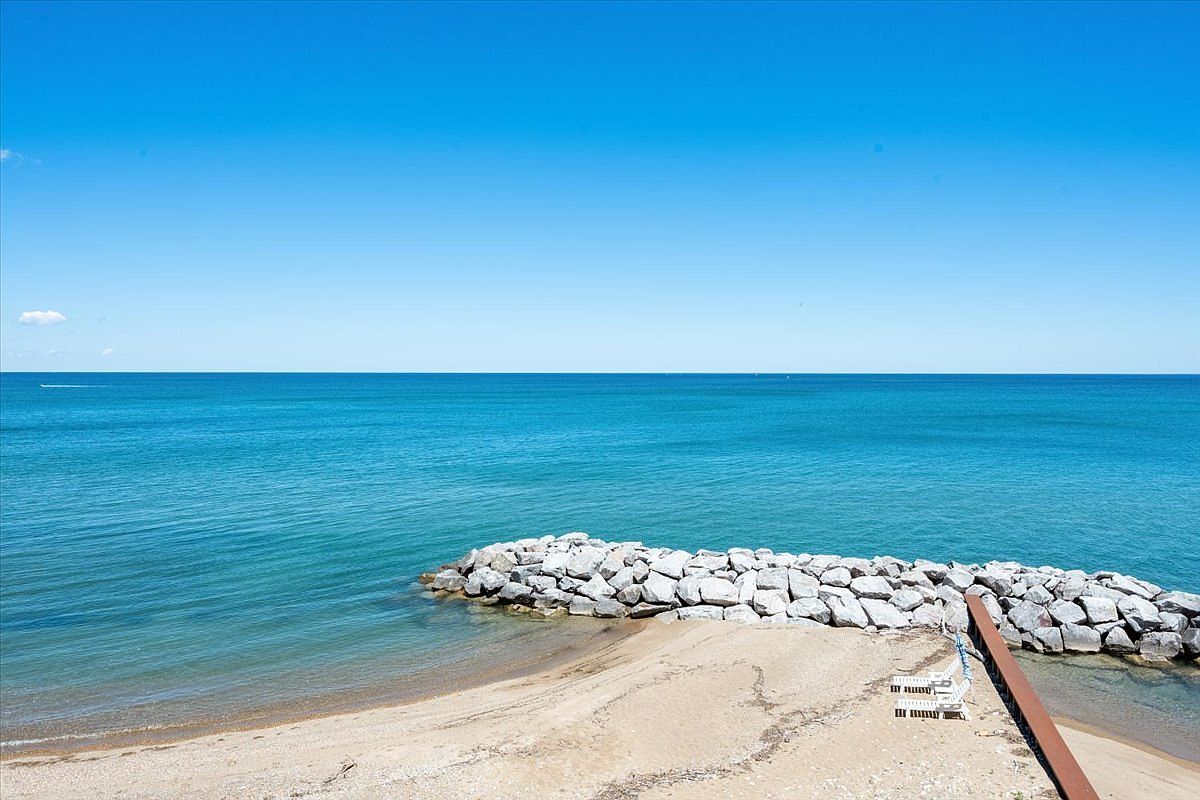 This elevated perspective captures a serene beachfront scene featuring a sandy shoreline meeting the vast, calm blue water. A prominent stone breakwater extends into the sea, providing a textured contrast to the smooth horizon and clear sky. Two white lounge chairs are positioned on the sand near a wooden structure, suggesting a private and tranquil outdoor retreat.