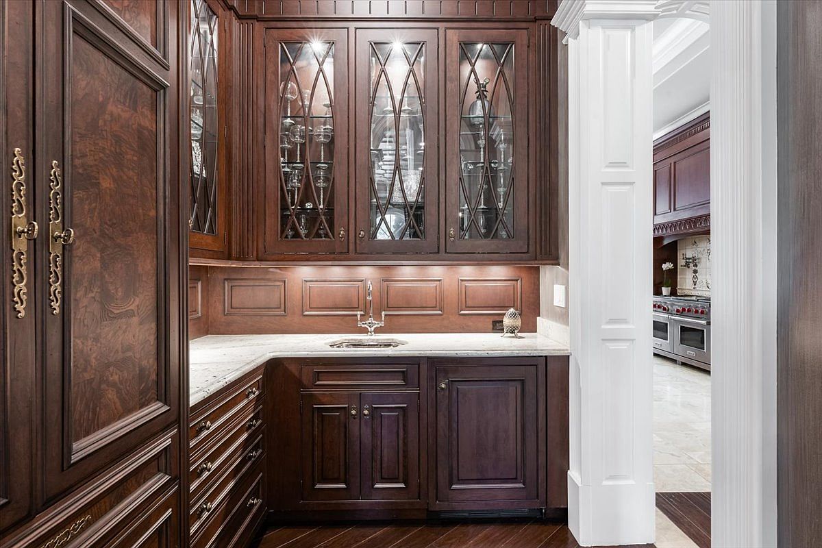 This elegant butler's pantry features rich, dark wood cabinetry with glass-front upper doors and intricate metal hardware. A light-toned stone countertop provides a striking contrast to the cabinetry, complemented by an under-cabinet lighting system and a small prep sink. The perspective looks directly into the wet bar area, with a glimpse of a larger kitchen space visible through a white-trimmed doorway in the background.
