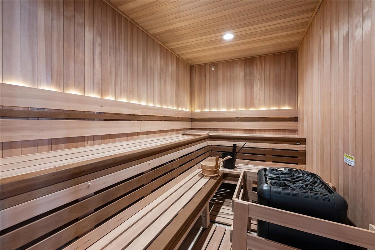This image showcases a professionally designed sauna room featuring warm-toned wood paneling throughout the walls and tiered bench seating. A black electric sauna heater filled with dark stones sits in the corner, accompanied by a wooden bucket and ladle. The space is illuminated by subtle, integrated strip lighting that creates a relaxing and inviting atmosphere.