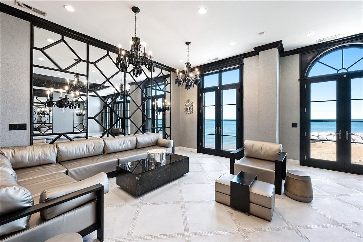 This sophisticated living room features a large, metallic-toned sectional sofa and a matching armchair arranged around a dark, polished stone coffee table. The space is defined by a dramatic, geometric mirrored wall and two ornate black chandeliers, while tall glass doors provide expansive views of the ocean and shoreline. The light-colored tiled flooring with decorative inlays enhances the luxurious, modern aesthetic of the room.