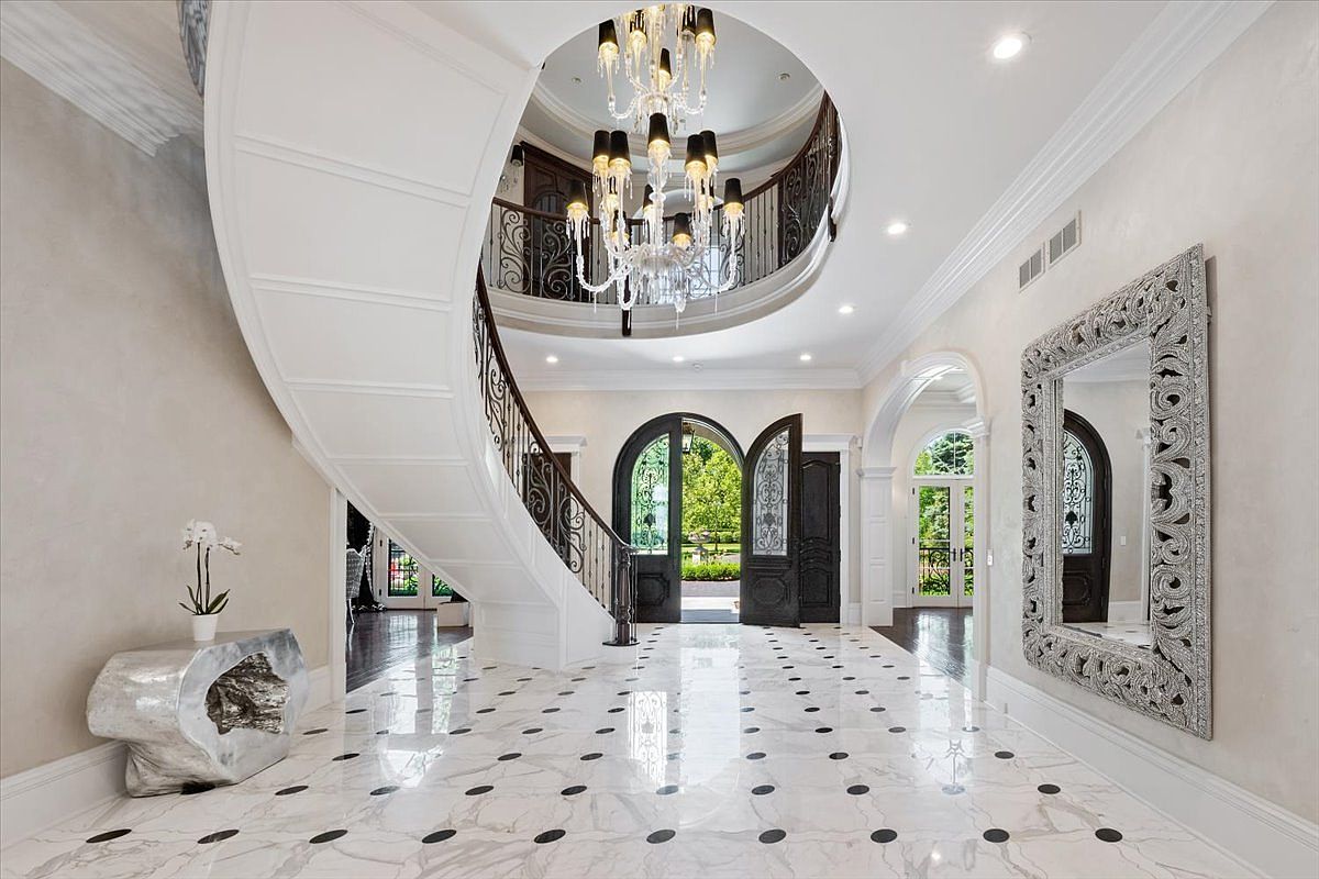 This grand foyer features a sweeping curved staircase with intricate metal railings that leads to an upper landing. The space is anchored by a large crystal chandelier suspended from a circular ceiling cutout, illuminating the polished stone flooring with dark circular inlays. A large, ornate silver-framed mirror hangs on the wall, reflecting the bright, open entryway that leads through double doors to the outdoors.