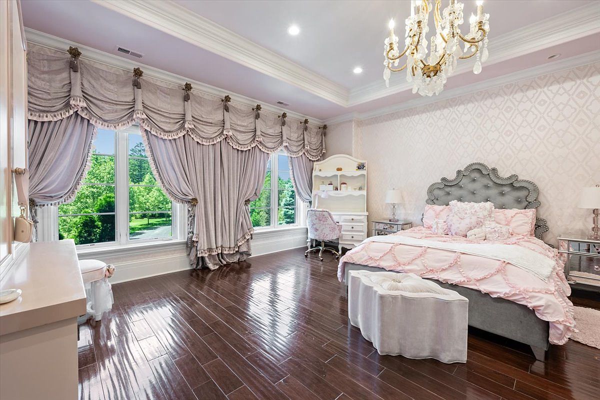 This elegant bedroom features a sophisticated design with a tufted gray headboard, a plush pink ruffled bedspread, and a matching upholstered bench at the foot of the bed. The room is accented by ornate, heavy drapery over large windows, a crystal chandelier, and patterned wallpaper, all set against dark wood flooring. A white desk and chair are tucked into the corner, creating a refined and feminine aesthetic.