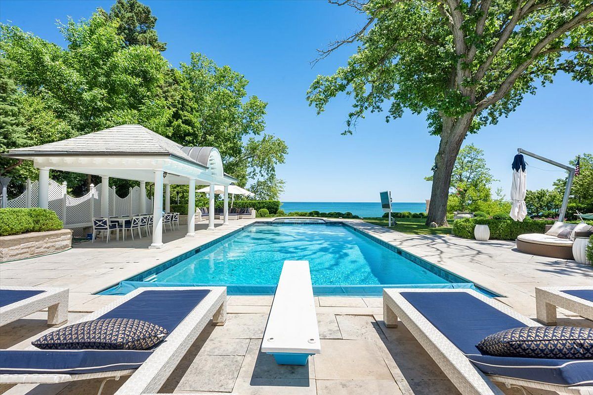 This luxurious outdoor space features a long, rectangular swimming pool with a diving board, set against a backdrop of lush trees and a clear blue sky. To the left, a covered pavilion with white columns provides a shaded seating area, while lounge chairs are arranged on the stone patio surrounding the pool. The perspective looks directly down the length of the pool toward the horizon, emphasizing the serene, resort-like atmosphere of the property.