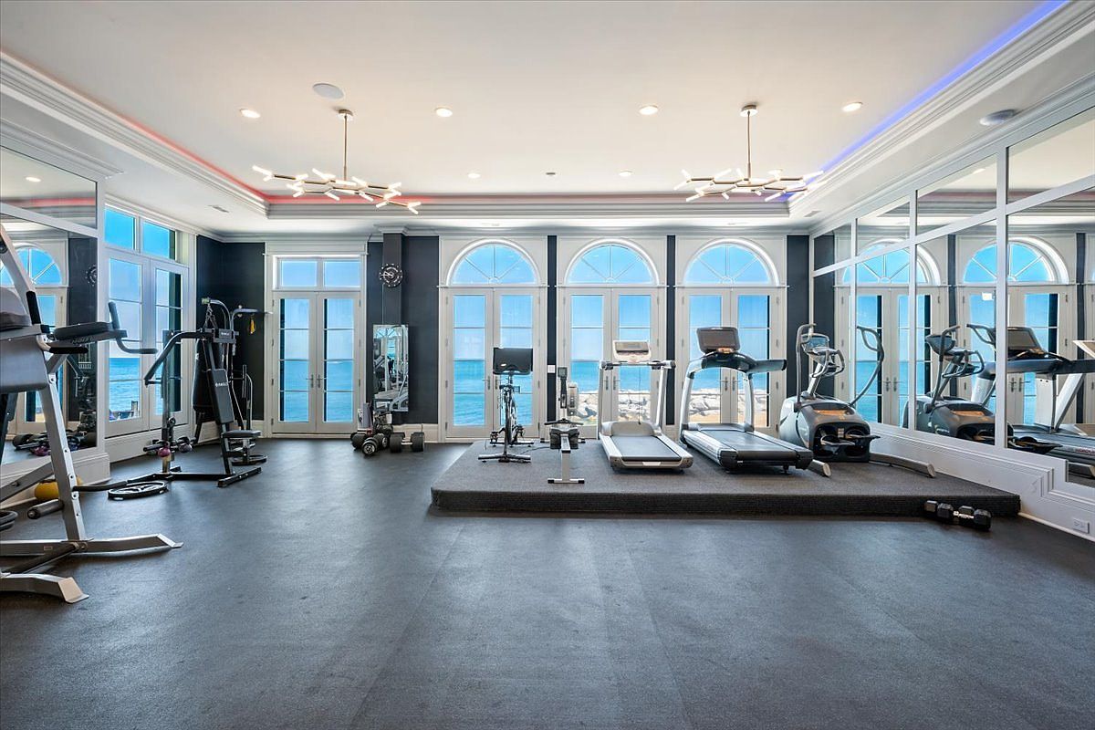 This spacious home gym features a dark rubberized floor and a raised platform housing a treadmill, elliptical, and stationary bike. Large, arched glass doors provide an expansive view of the ocean, while mirrored walls on either side enhance the sense of depth and brightness. The room is illuminated by modern, multi-armed light fixtures and recessed lighting, creating a clean and professional workout environment.