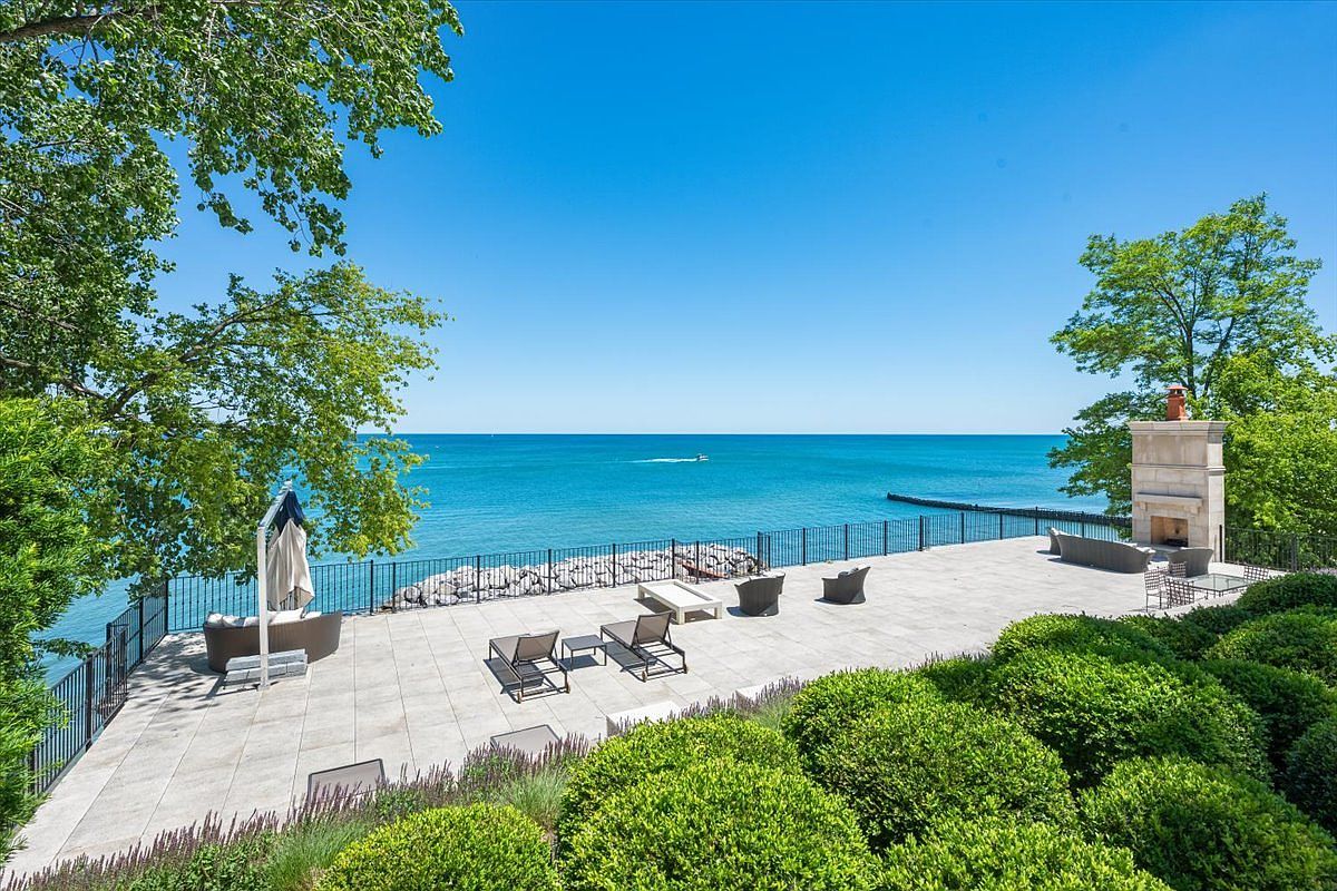 This expansive stone-paved patio offers a stunning, elevated vantage point overlooking a vast, calm body of water. The space is furnished with multiple lounge chairs and a seating area centered around a prominent stone-clad chimney breast, all framed by lush green landscaping and a black metal railing. The clear blue sky and open horizon create a serene, high-end atmosphere perfect for outdoor entertaining.