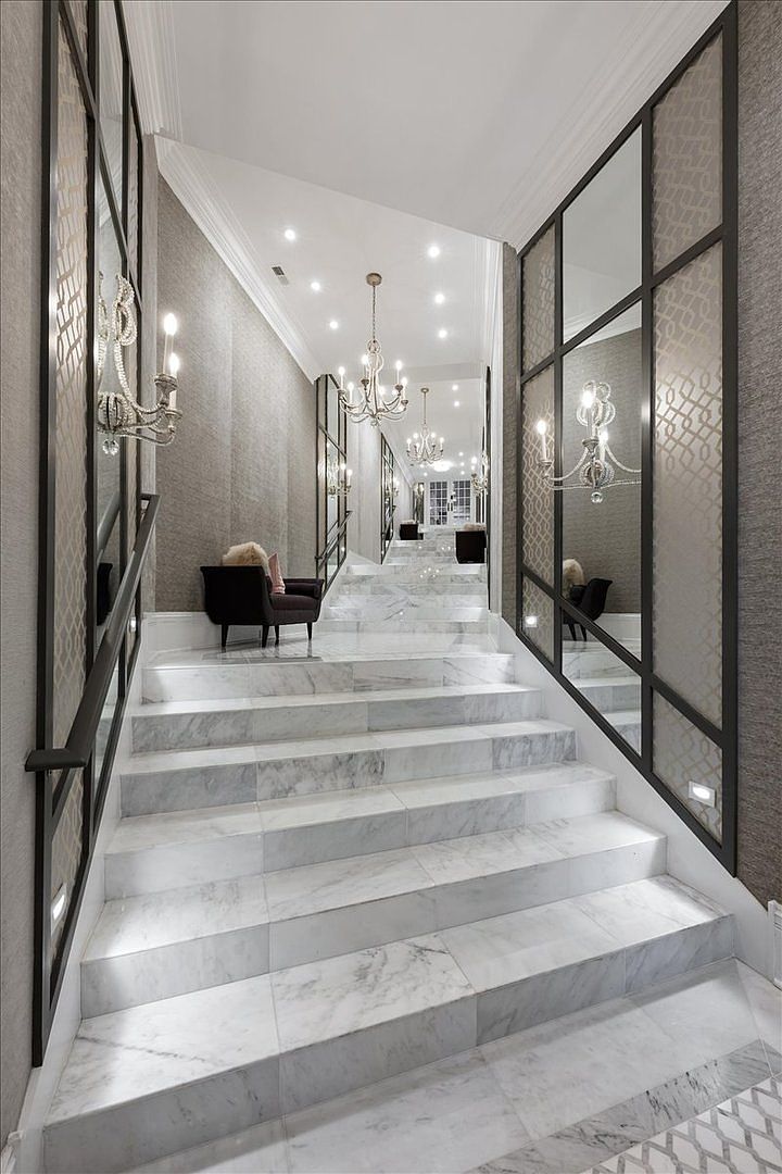 This grand staircase features wide stone steps leading upward through a formal entryway. The walls are adorned with large, grid-patterned mirrors and decorative wall sconces, creating a symmetrical and sophisticated aesthetic. A dark accent chair sits on the landing, while multiple chandeliers provide elegant overhead lighting, emphasizing the luxurious and spacious feel of the home.