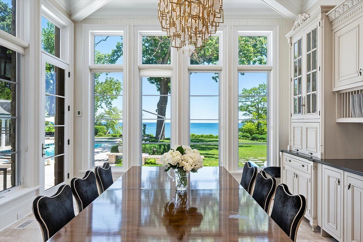 This elegant dining room features a long, polished wood table surrounded by dark upholstered chairs, centered beneath a sophisticated gold-toned chandelier. A wall of tall windows provides an expansive, unobstructed view of a lush green lawn, a swimming pool, and the open water beyond. To the right, a built-in white hutch with glass-front cabinetry adds a classic, refined touch to the bright and airy space.