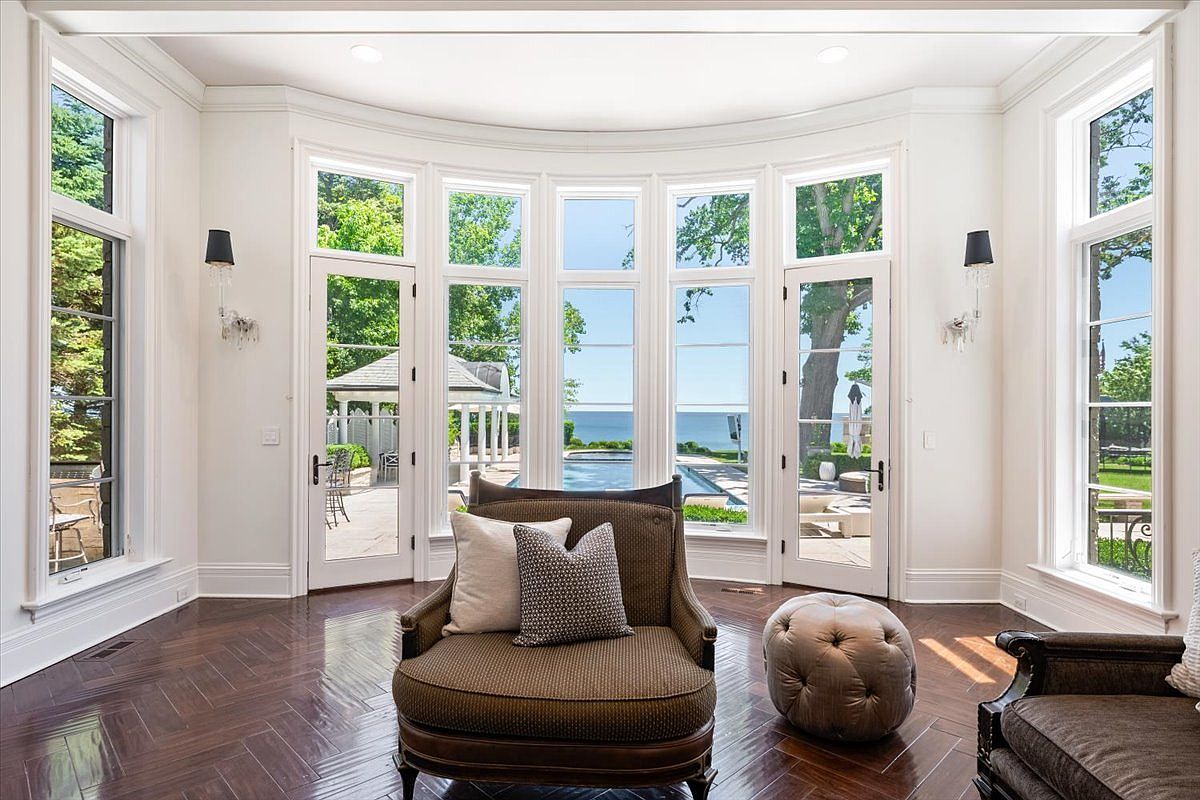 This bright sunroom features a curved wall of large windows and glass doors that provide expansive views of the pool and the water beyond. The space is anchored by dark, herringbone-patterned wood flooring and includes a comfortable armchair with patterned pillows and a tufted ottoman. Symmetrical wall sconces flank the windows, creating an elegant and serene atmosphere perfect for relaxation.