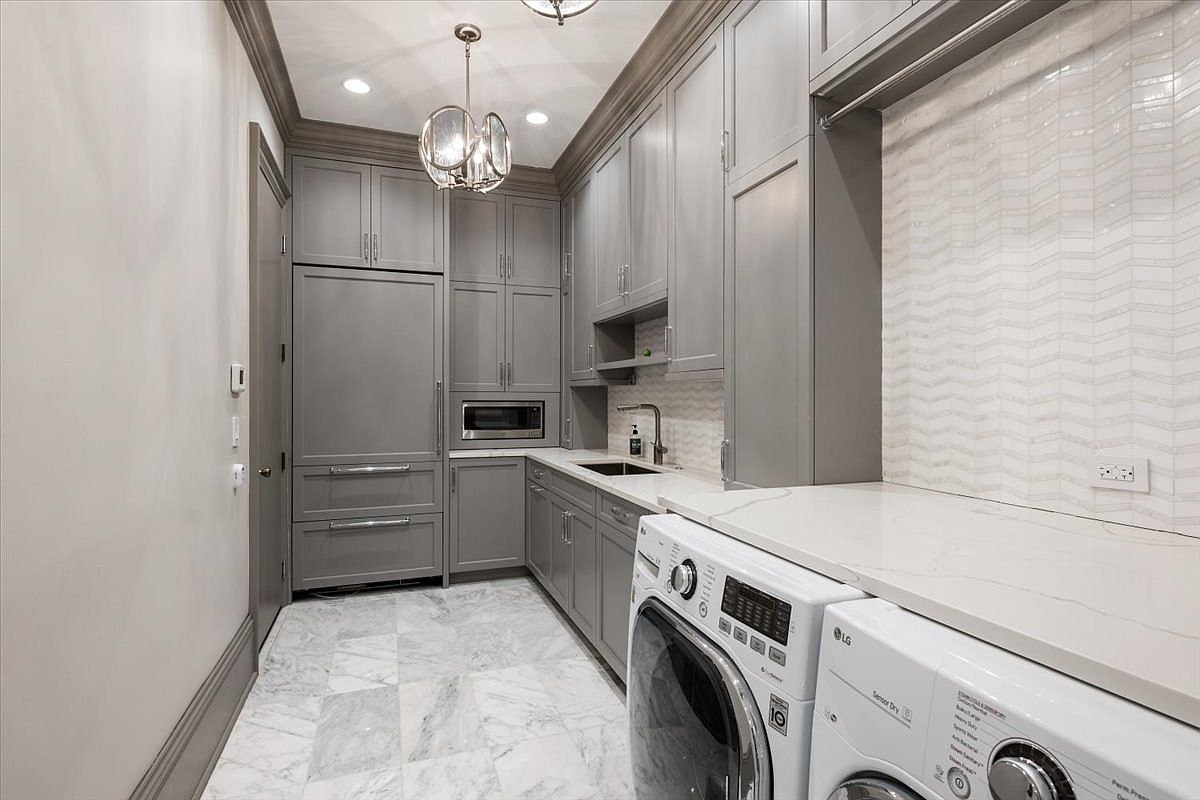 This sophisticated laundry room features sleek grey cabinetry, a built-in refrigerator, and a microwave, blending utility with high-end design. The space is finished with a patterned stone floor and a light-colored chevron-patterned backsplash, complemented by a modern pendant light fixture. The perspective captures the full length of the room, showcasing the integrated washer and dryer set beneath a clean, light-colored countertop.