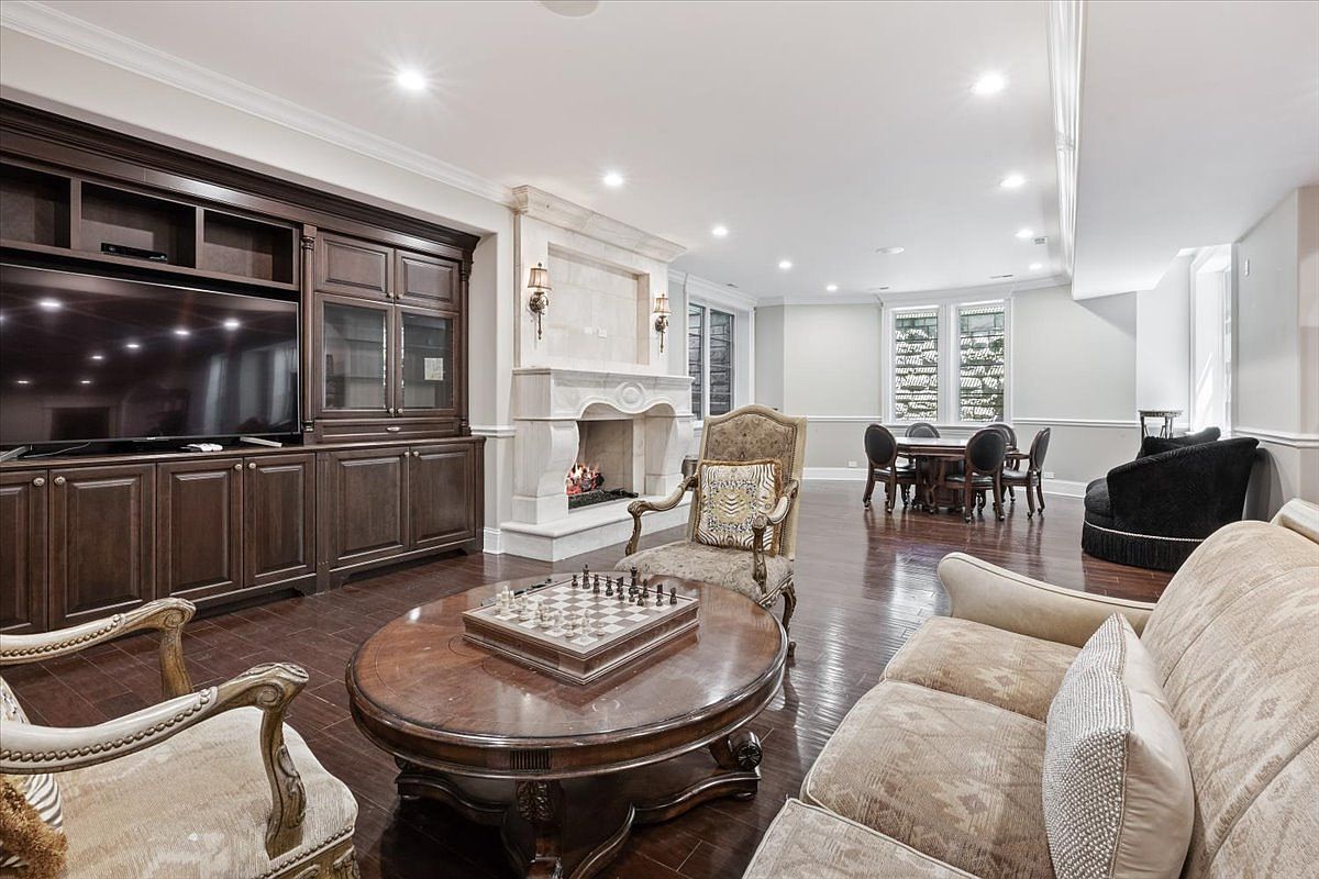 This spacious living room features dark wood flooring and a large, ornate stone fireplace surround that serves as a central focal point. A built-in dark wood entertainment center spans the wall, providing ample storage and display space, while a chess set sits atop a round wooden coffee table in the foreground. The room transitions into a dining area in the background, illuminated by recessed lighting and natural light from multiple windows.