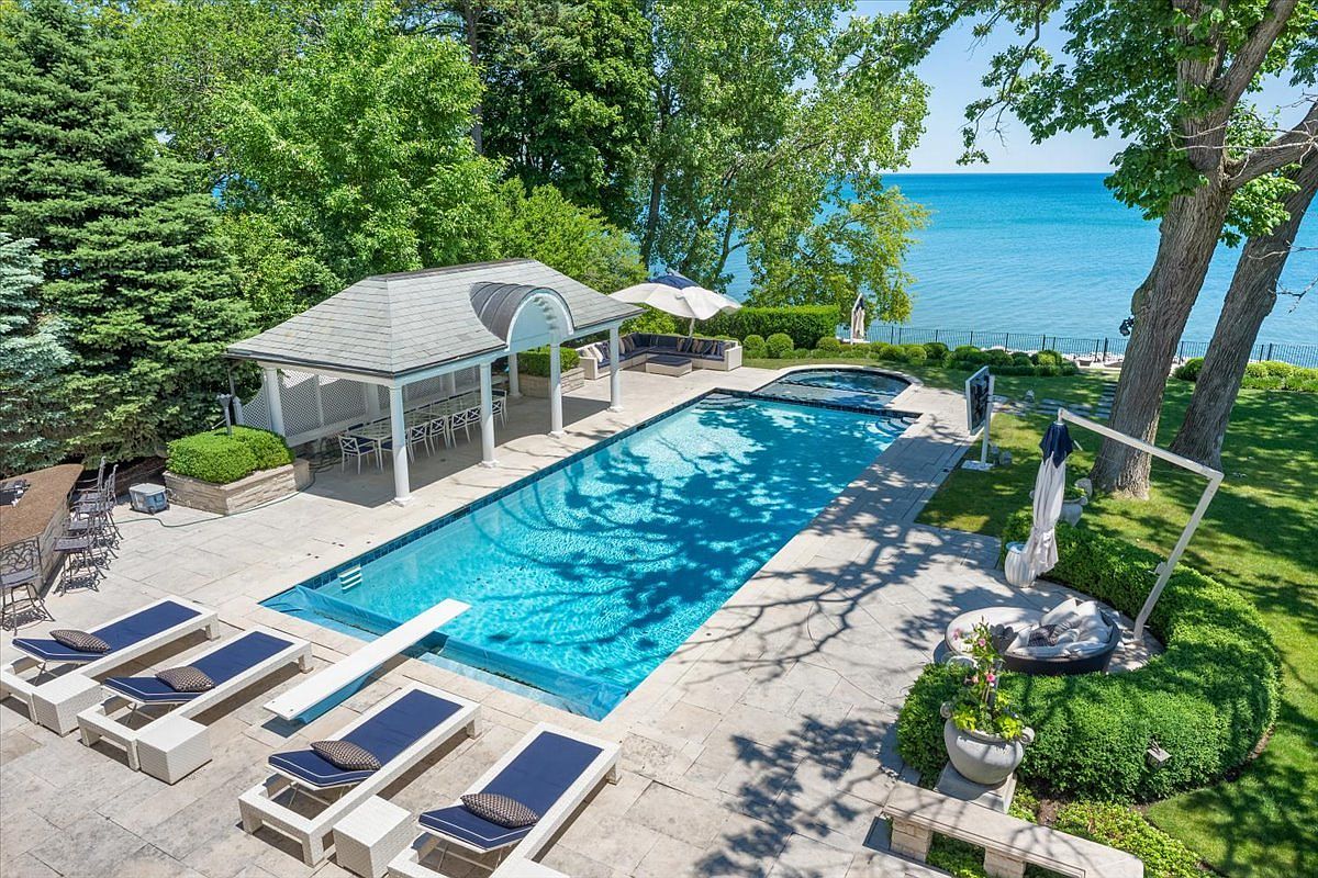 This high-angle aerial view captures a luxurious backyard oasis featuring a rectangular swimming pool with an integrated spa overlooking a vast body of water. The stone-paved patio is furnished with multiple lounge chairs, a covered pavilion with dining space, and a circular seating area nestled near mature trees. The lush green lawn and expansive lake view create a serene and high-end atmosphere perfect for outdoor entertaining.