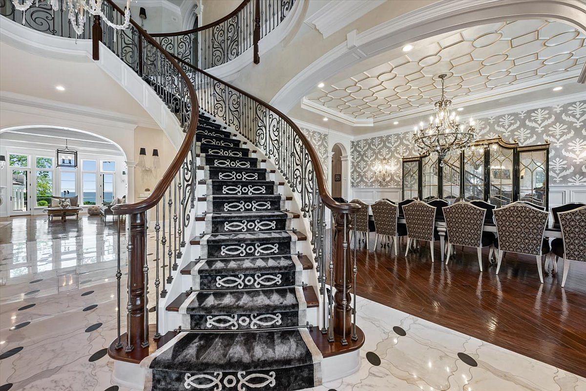 This grand foyer features a sweeping, curved staircase with intricate wrought-iron railings and a patterned runner that serves as the central focal point. To the right, a formal dining area is visible, characterized by ornate wallpaper, a large crystal chandelier, and a coffered ceiling with geometric detailing. The space is finished with polished stone flooring, creating a luxurious and expansive atmosphere that flows seamlessly into the adjacent rooms.