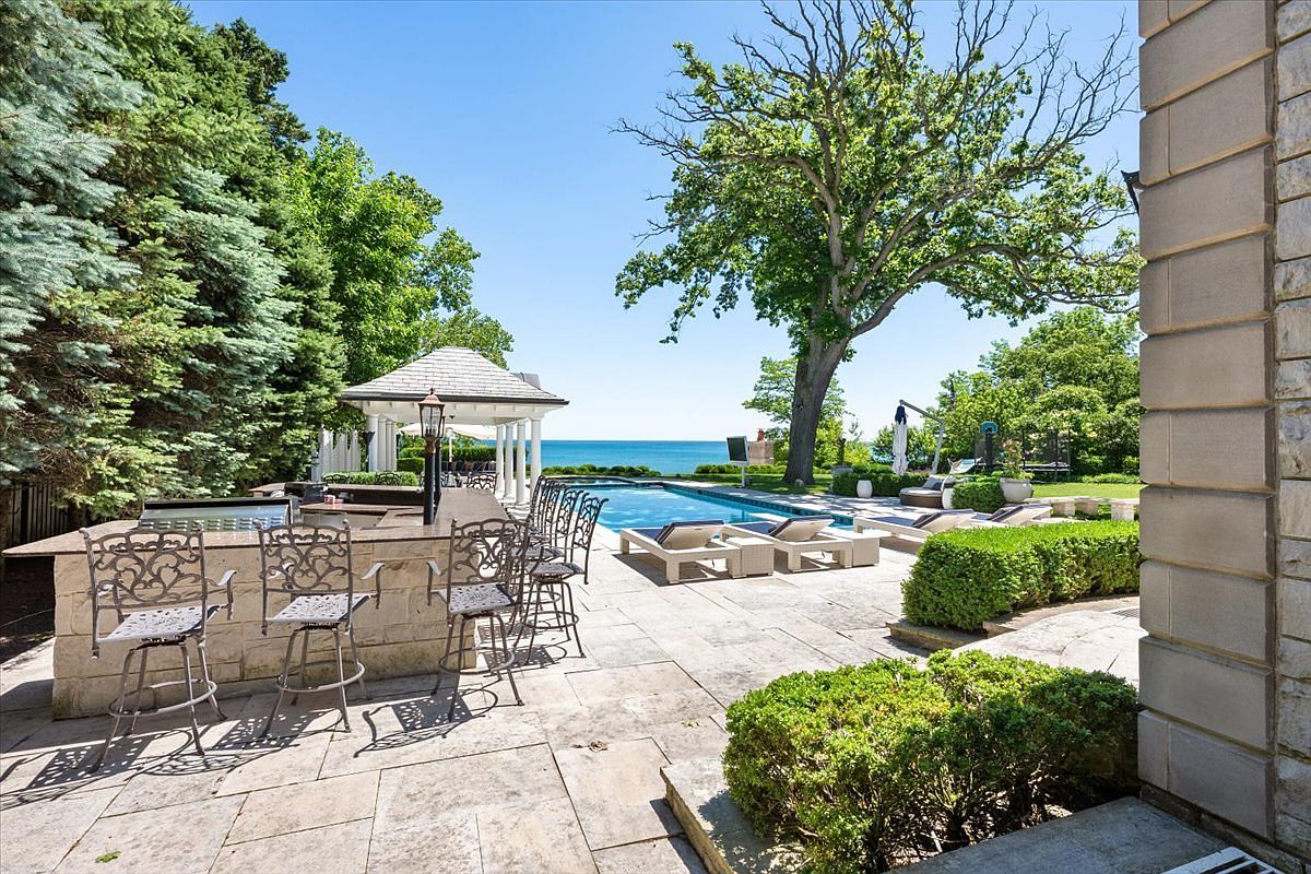 This expansive outdoor living space features a stone-paved patio with a built-in bar area, complete with high-backed metal stools and a grill station. A rectangular swimming pool serves as the centerpiece, leading the eye toward a scenic waterfront view framed by mature trees and a gazebo. The setting offers a luxurious, resort-like atmosphere perfect for entertaining and relaxation.