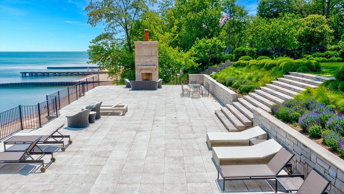 This expansive stone-paved patio offers a luxurious outdoor living space overlooking a serene lakefront. The area features a prominent stone chimney breast with an integrated fireplace, multiple lounge chairs, and a dining set, all framed by tiered landscaping and stone retaining walls. The perspective captures the seamless transition from the manicured terrace to the natural beauty of the water and shoreline below.