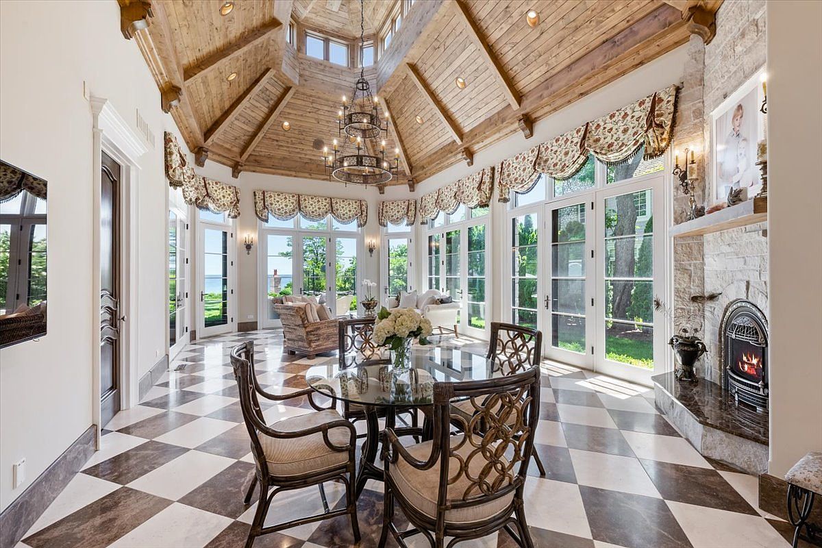 This elegant sunroom features a dramatic vaulted wood-paneled ceiling with exposed beams and a central chandelier. The space is anchored by a stone-clad chimney breast with a built-in fireplace and is finished with a classic diamond-patterned tile floor. Large windows and glass doors provide abundant natural light and views of the surrounding landscape, creating a bright and sophisticated atmosphere.