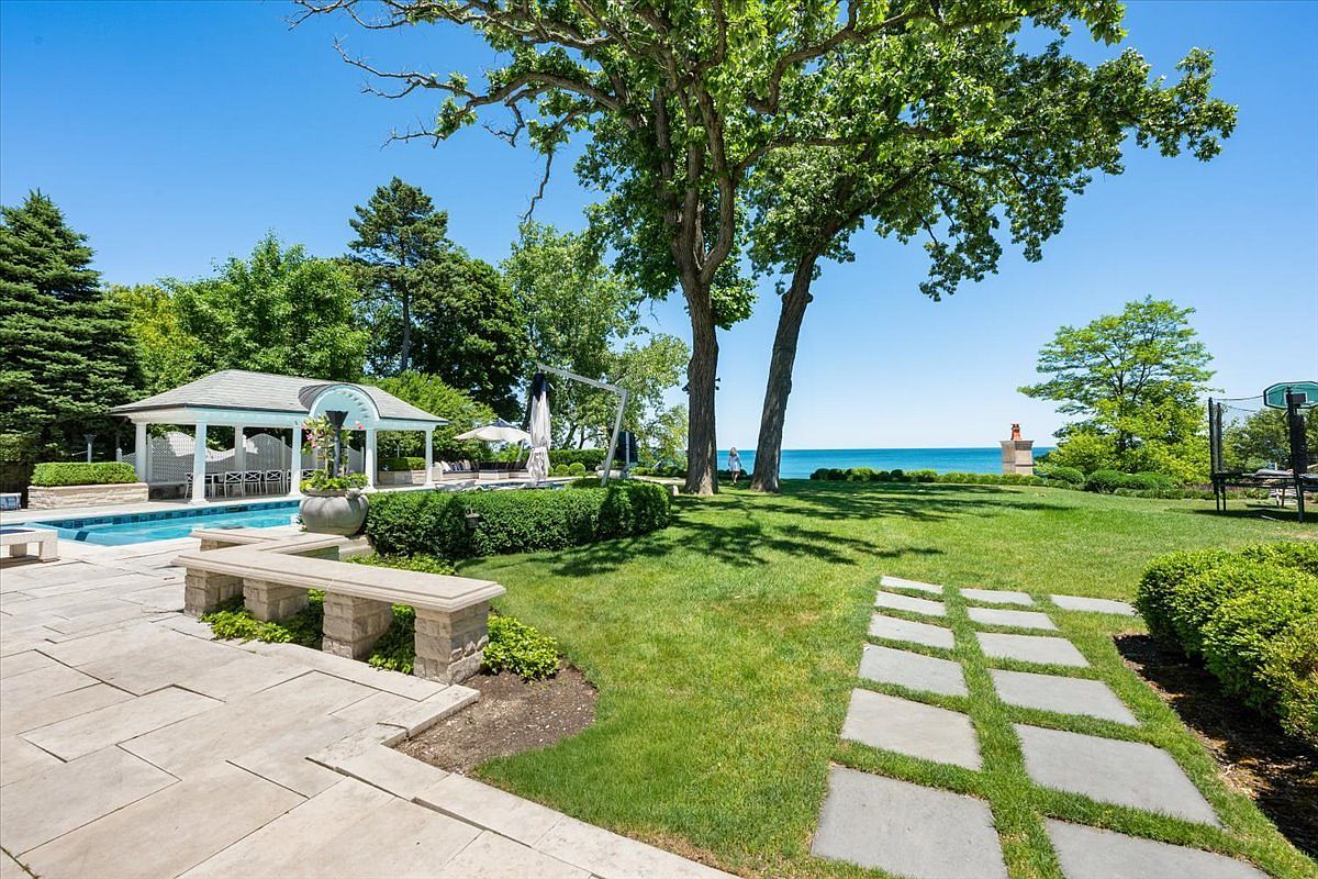 This expansive outdoor living space features a pristine swimming pool adjacent to a classic white gazebo, set against a backdrop of lush green lawn and mature trees. A stone-paved patio provides a transition to the grass, where a series of rectangular stepping stones leads the eye toward the scenic waterfront view in the distance. The scene captures a serene, resort-like atmosphere perfect for relaxation and outdoor entertaining.