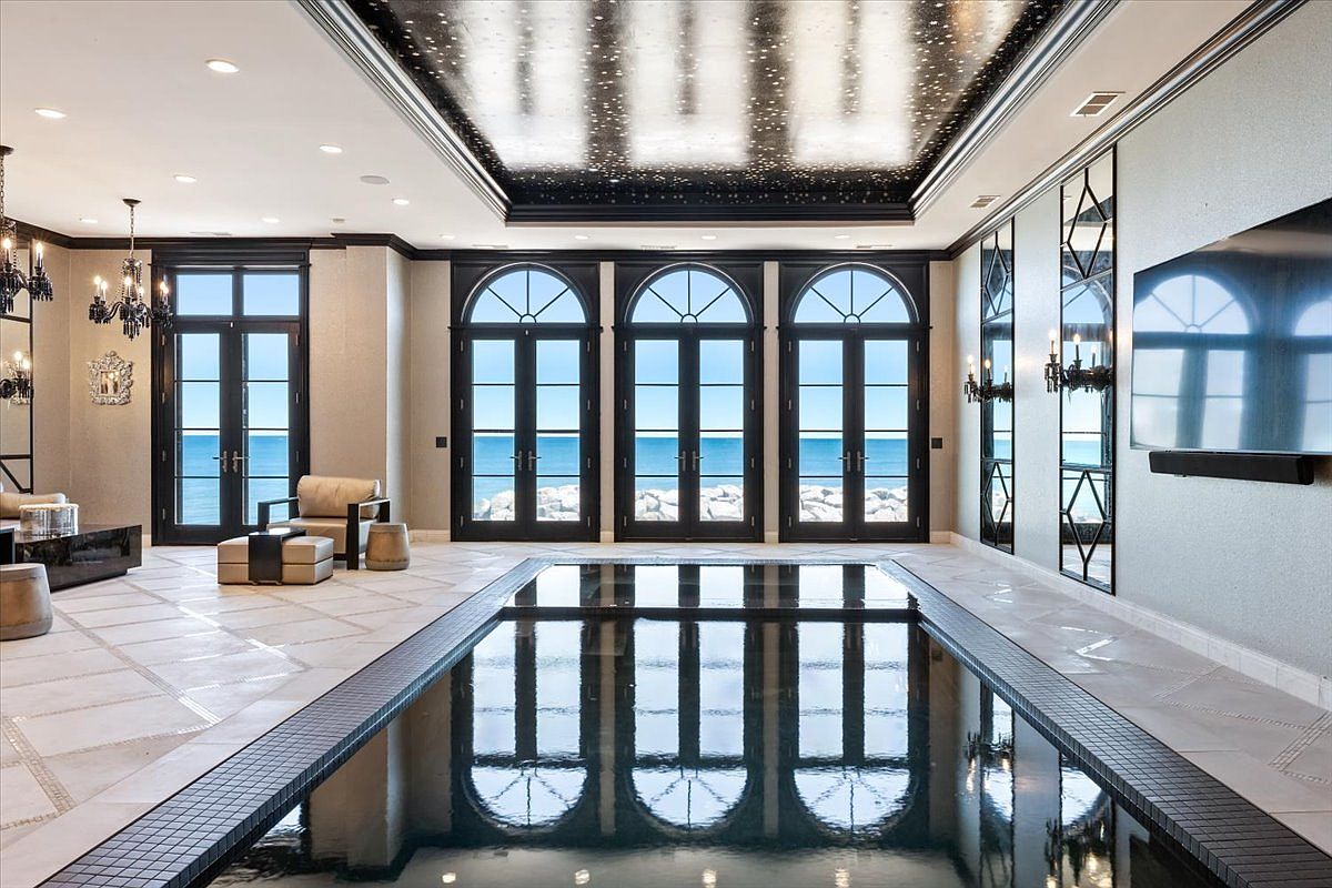 This luxurious indoor pool room features a dark, reflective pool surface that mirrors the three sets of arched glass doors leading to a waterfront view. The ceiling is a dramatic, dark-toned feature with a starlit effect, complemented by elegant chandeliers and mirrored wall panels. The space is furnished with comfortable seating, creating a sophisticated and serene atmosphere for relaxation.