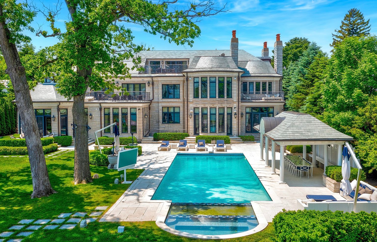 This expansive rear view of a grand stone estate showcases a symmetrical architectural design with multiple levels of tall windows and wrought-iron balconies. The meticulously landscaped backyard features a large rectangular swimming pool with an integrated spa, surrounded by a stone patio and a covered outdoor dining pavilion. The scene is framed by lush green trees and manicured hedges, creating a private and luxurious outdoor living environment.