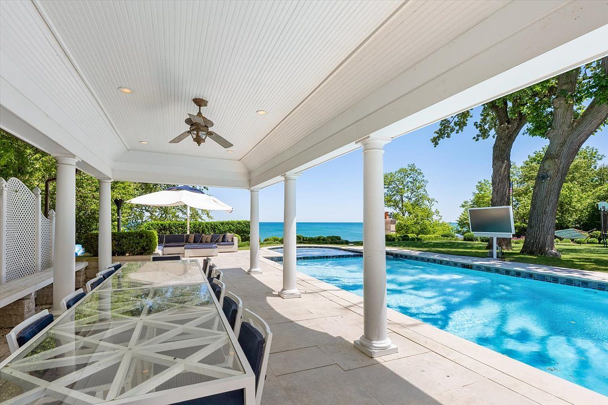 This expansive covered patio offers a luxurious outdoor living space, featuring a long glass-topped dining table and a comfortable lounge area shaded by a white umbrella. The space seamlessly transitions to a sparkling swimming pool that overlooks a scenic waterfront view, framed by lush green trees. The architectural design includes classic white columns and a beadboard ceiling, creating an elegant and inviting atmosphere for relaxation or entertaining.
